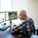 Landwirt im karierten Hemd sitzt vor 2 Bildschirmen im Büro